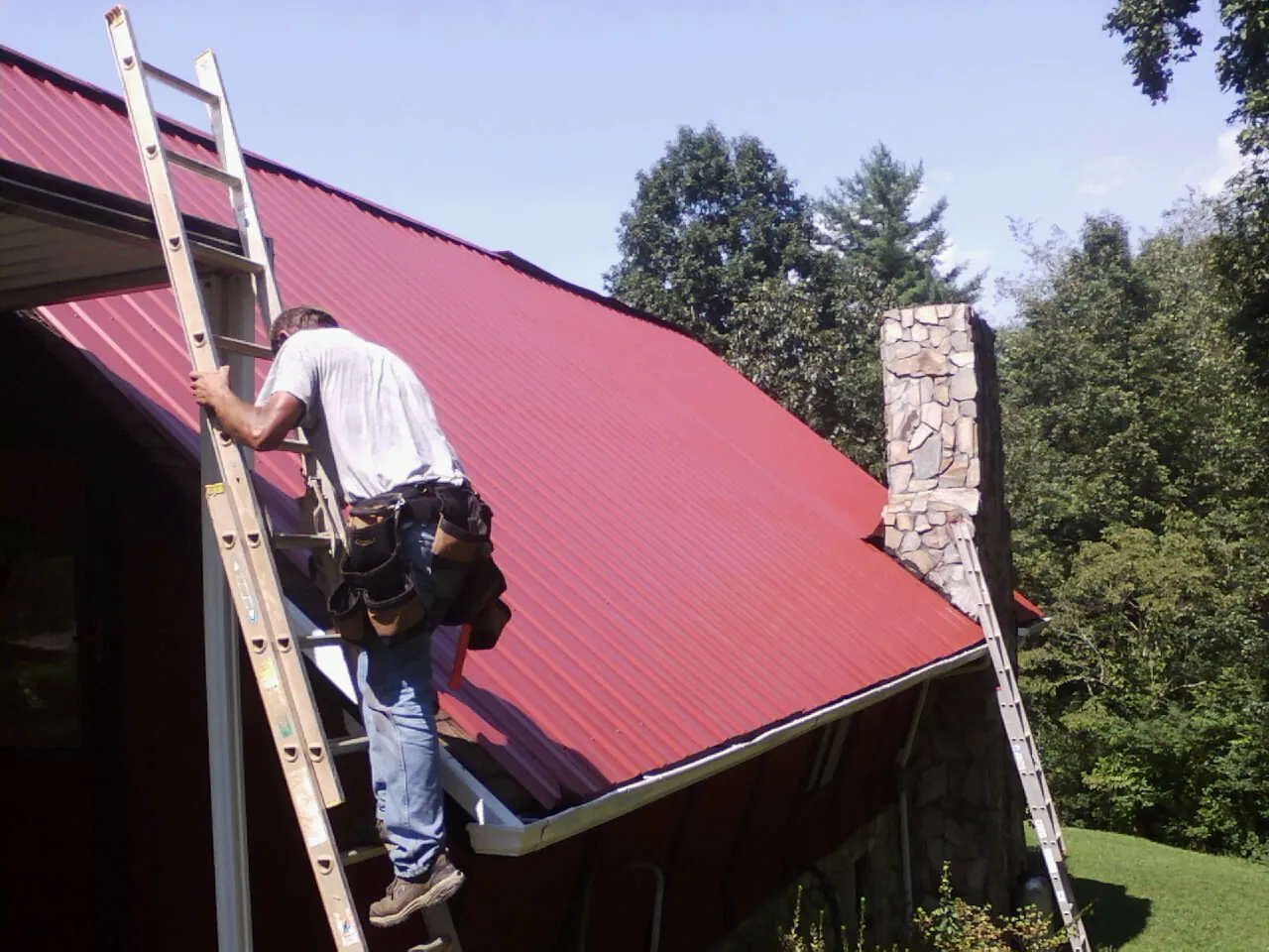 Roofer on a ladder beside a red metal roof and stone chimney — Soffit & Fascia Repair by Roofing Brothers in Southport